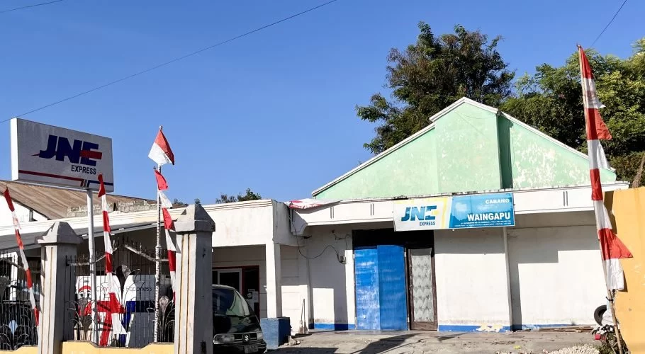 The exterior of the JNE Express Waingapu building features white walls, a light green roof, and blue signage, adorned with red and white Indonesian flags under a clear blue sky.