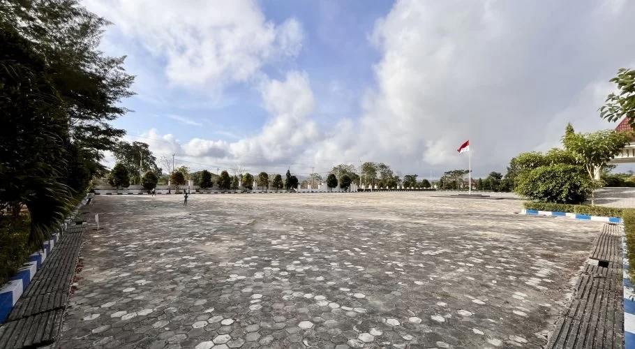 An expansive paved public area, popular for jogging, is lined with trees and shrubs under a cloudy blue sky, featuring an Indonesian flag.