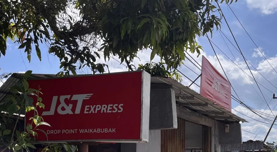 A red J&T Express drop point sign in Waikabubak, Sumba, is partially visible beneath green tree leaves against a bright blue sky.