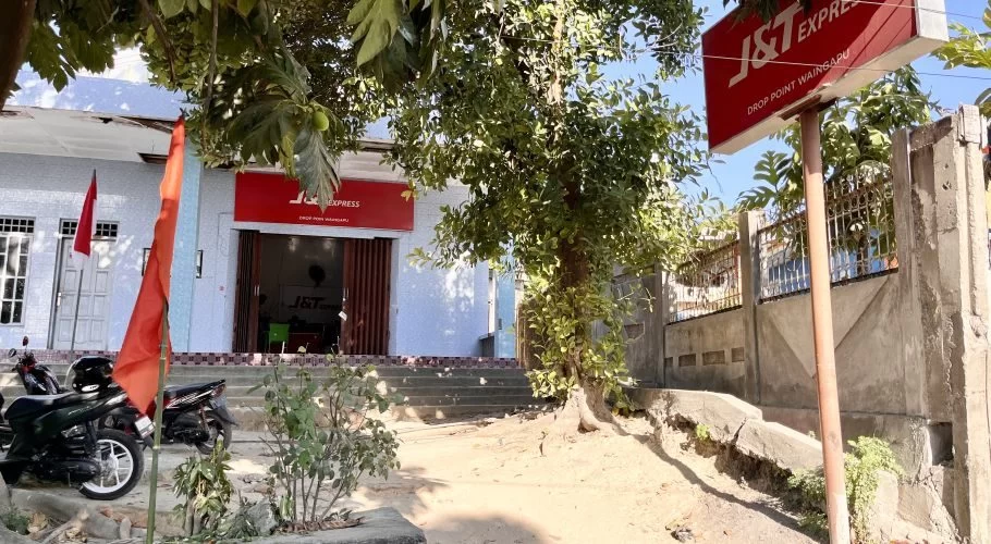 The J&T Express Waingapu branch building with its prominent red sign, two motorcycles, and a red flag under a tree on a sunny day.