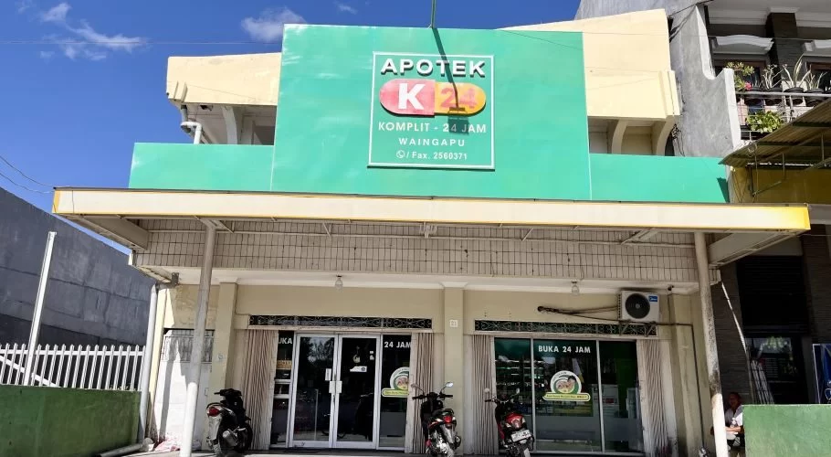 The exterior of K-24 Pharmacy in Waingapu features a prominent green sign, a light-colored facade, and two motorcycles parked below, under a blue sky.