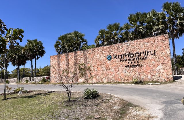 The large, textured stone entrance sign for Kambaniru Beach Hotel & Resort, with black lettering and a blue logo, stands under a clear blue sky amidst palm trees.