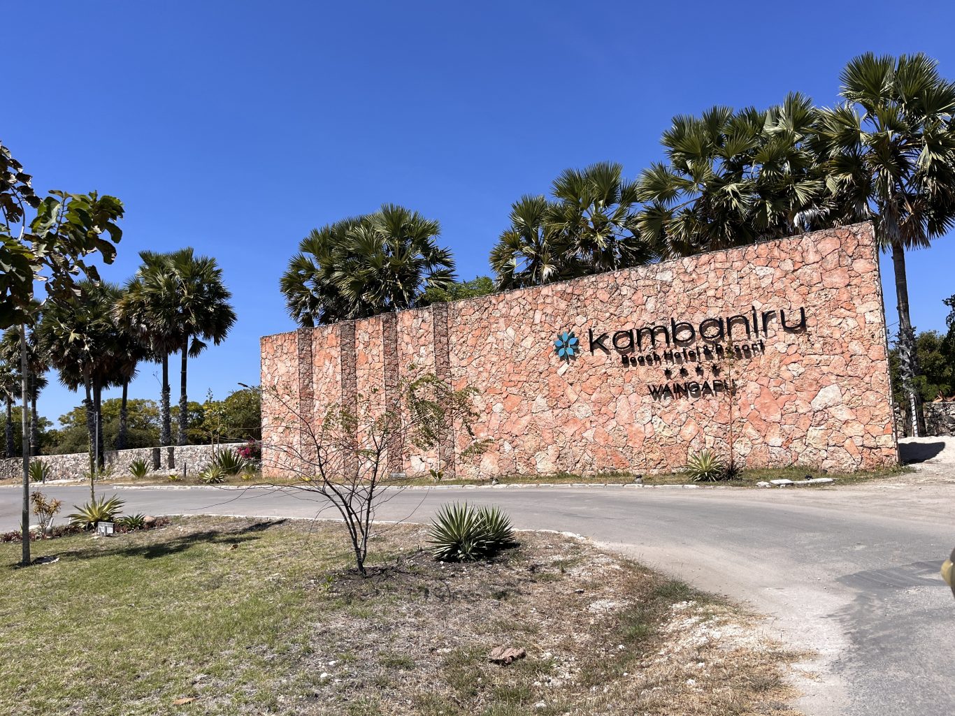The large, textured stone entrance sign for Kambaniru Beach Hotel & Resort, with black lettering and a blue logo, stands under a clear blue sky amidst palm trees.