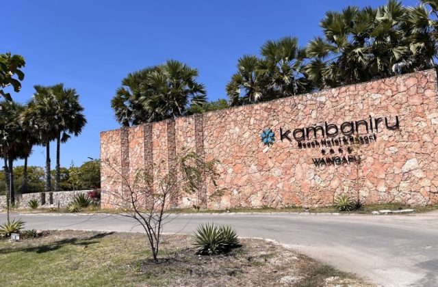 The large, textured stone entrance sign for Kambaniru Beach Hotel & Resort, with black lettering and a blue logo, stands under a clear blue sky amidst palm trees.