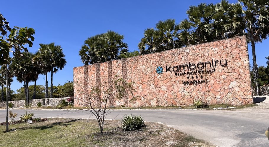 The large, textured stone entrance sign for Kambaniru Beach Hotel & Resort, with black lettering and a blue logo, stands under a clear blue sky amidst palm trees.