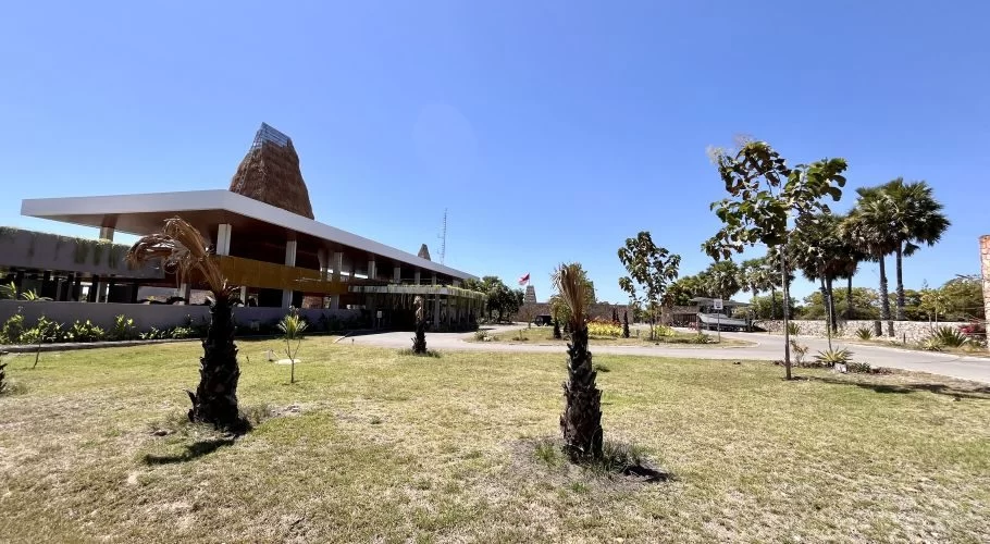The modern Kambaniru Beach Hotel and Resort building with a traditional thatched roof stands amidst expansive green grounds under a clear blue sky in East Sumba.