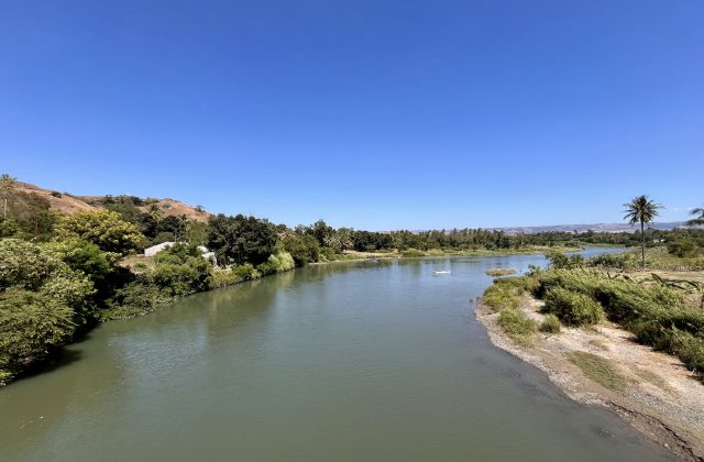 The wide, calm Kambaniru River flows through lush green banks with coconut trees and distant brown hills under a clear blue sky in East Sumba.
