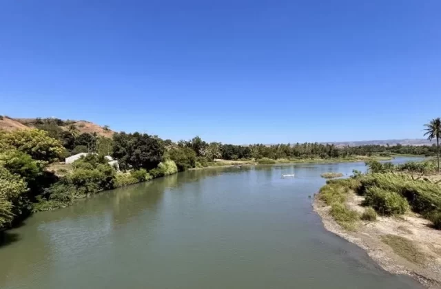 The wide, calm Kambaniru River flows through lush green banks with coconut trees and distant brown hills under a clear blue sky in East Sumba.