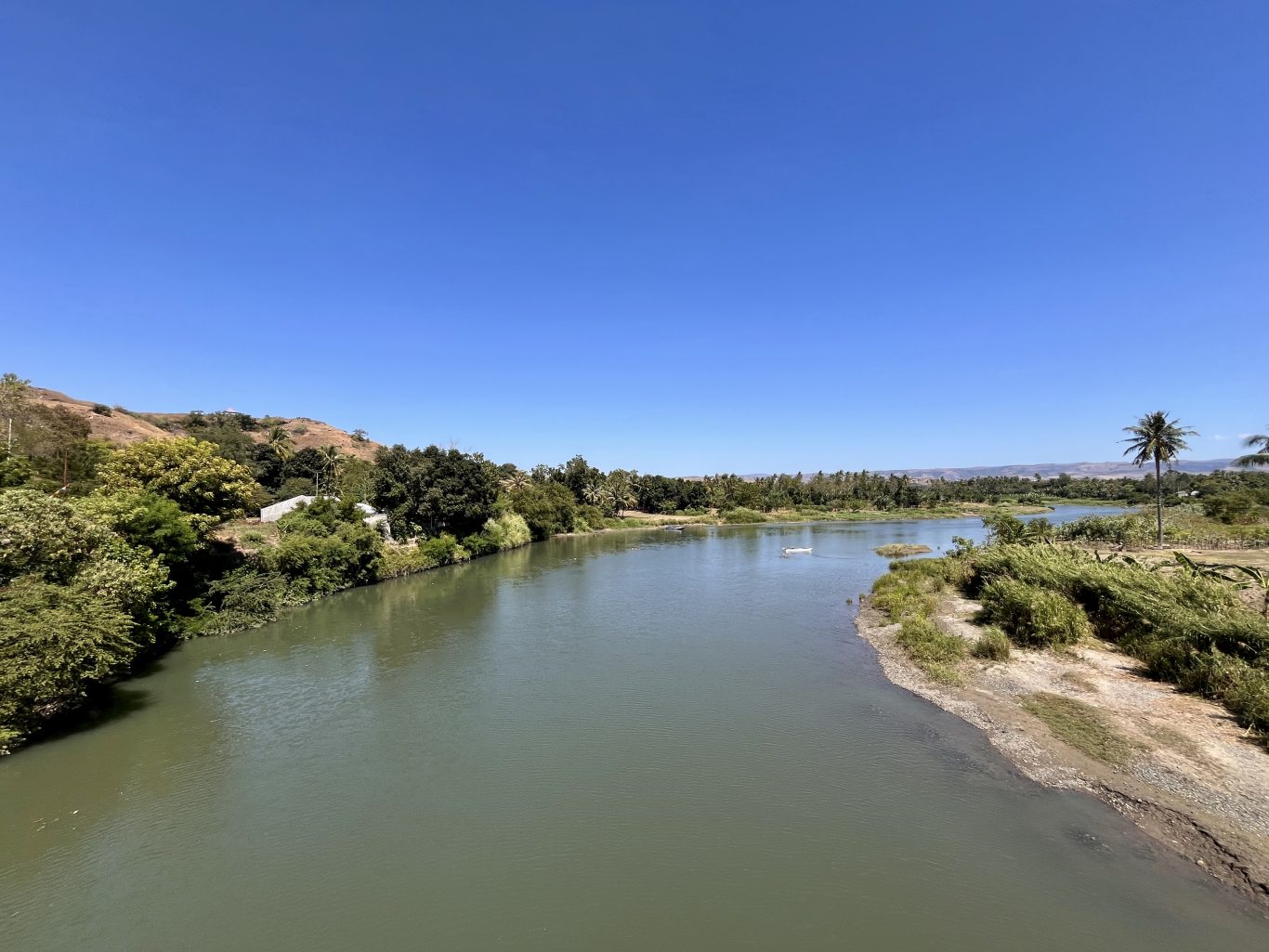 The wide, calm Kambaniru River flows through lush green banks with coconut trees and distant brown hills under a clear blue sky in East Sumba.