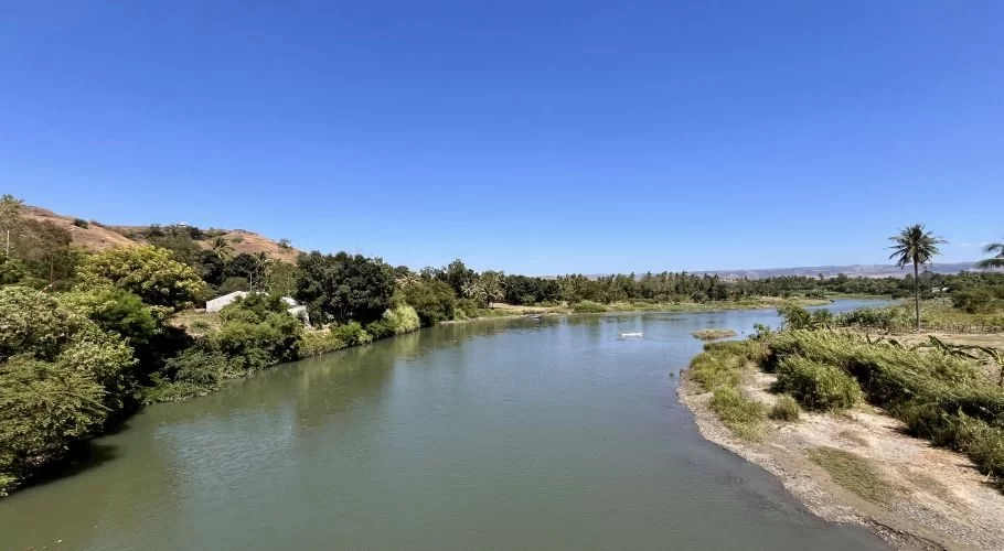 The wide, calm Kambaniru River flows through lush green banks with coconut trees and distant brown hills under a clear blue sky in East Sumba.