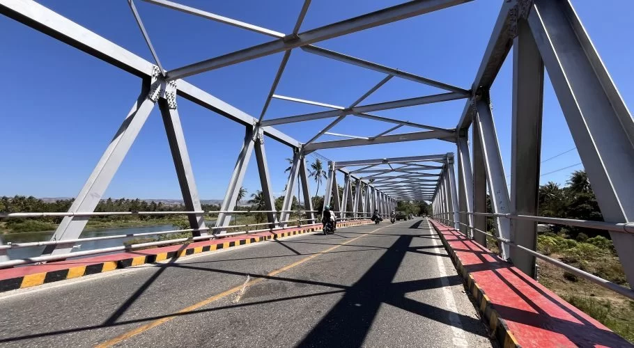 A grey metal truss bridge spans a blue-green river, with motorcycles driving on its paved road under a clear blue sky, flanked by lush green trees.