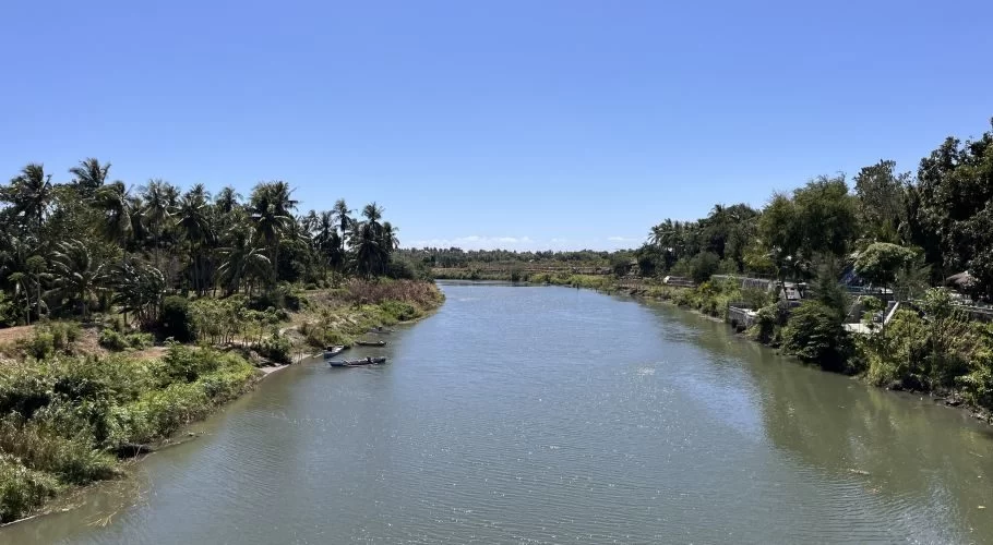 The wide Kambaniru River flows under a clear blue sky, flanked by dense green vegetation and numerous coconut trees, with small boats docked on the left bank.