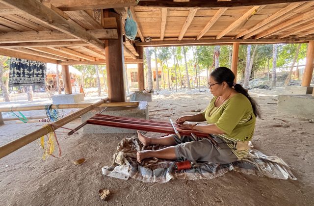 A woman in an olive shirt and grey pants sits barefoot on a mat, weaving red ikat textile on a traditional bamboo loom under a wooden stilt house.