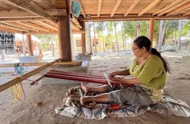 A woman in an olive shirt and grey pants sits barefoot on a mat, weaving red ikat textile on a traditional bamboo loom under a wooden stilt house.