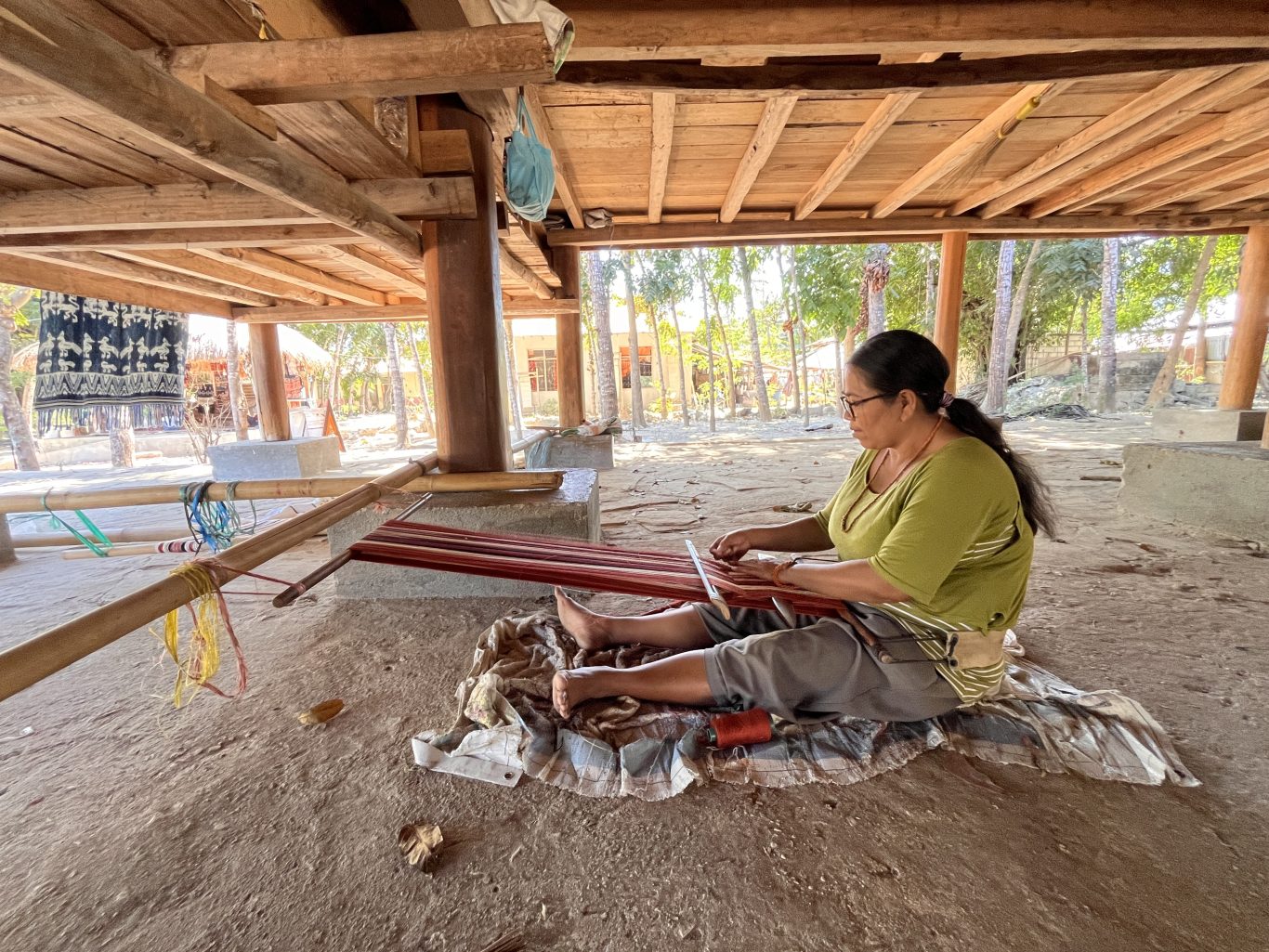 A woman in an olive shirt and grey pants sits barefoot on a mat, weaving red ikat textile on a traditional bamboo loom under a wooden stilt house.