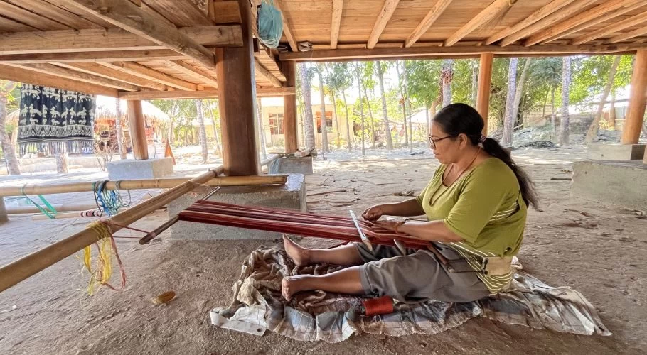 A woman in an olive shirt and grey pants sits barefoot on a mat, weaving red ikat textile on a traditional bamboo loom under a wooden stilt house.