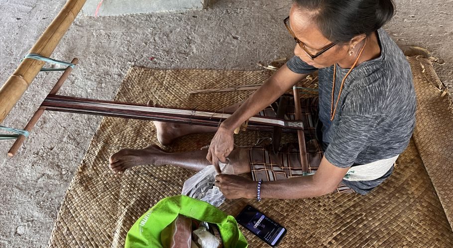 A Sumba woman with glasses sits on a woven mat, skillfully weaving colorful ikat threads on a traditional loom with simple tools.