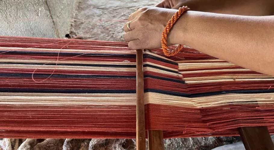 A person's hands with an orange beaded bracelet weave red, dark blue, and cream threads on a traditional loom.