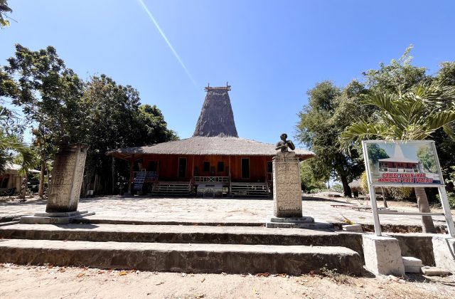 A traditional Sumba house with a tall thatched roof stands behind two carved stone megaliths and a gallery sign under a clear blue sky.