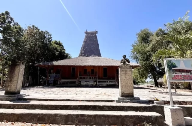 A traditional Sumba house with a tall thatched roof stands behind two carved stone megaliths and a gallery sign under a clear blue sky.