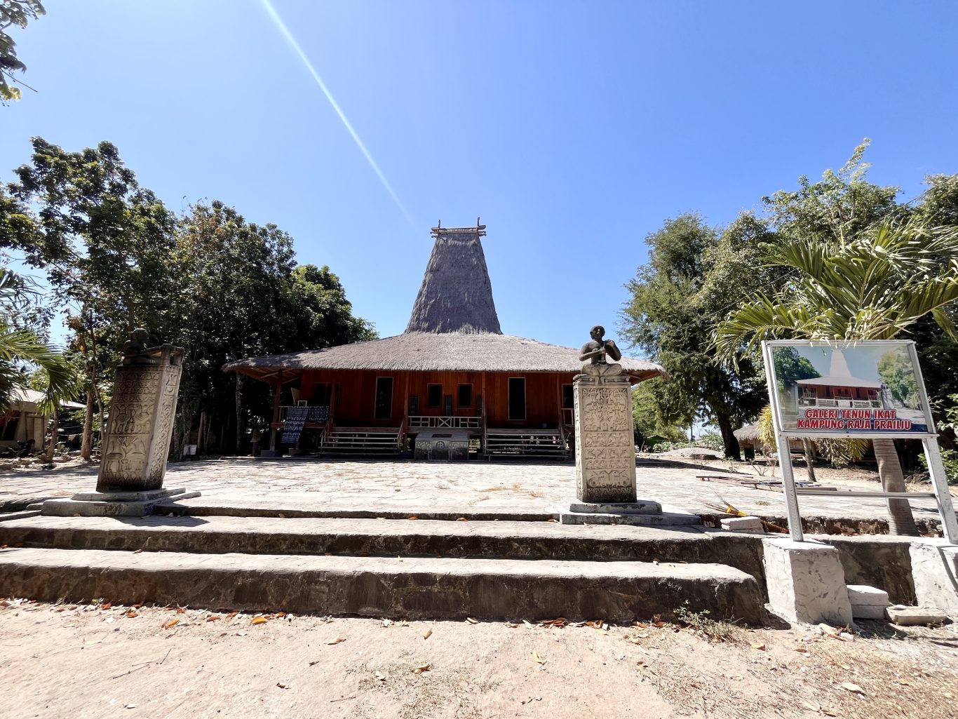 A traditional Sumba house with a tall thatched roof stands behind two carved stone megaliths and a gallery sign under a clear blue sky.