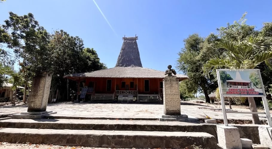 A traditional Sumba house with a tall thatched roof stands behind two carved stone megaliths and a gallery sign under a clear blue sky.