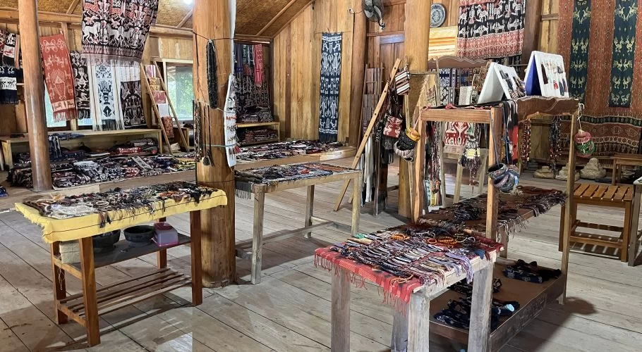 A wooden gallery interior showcases numerous colorful ikat textiles hanging on walls and draped over tables, alongside various handcrafted items.