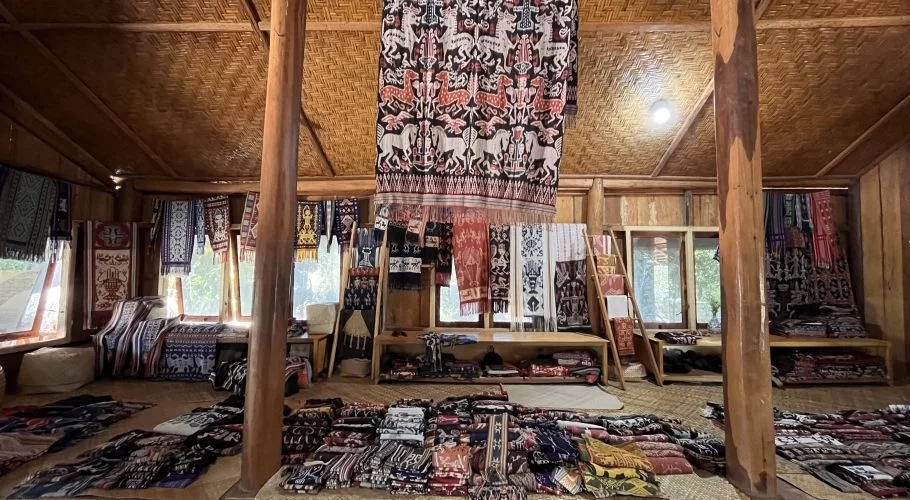 A traditional wooden gallery interior features numerous vibrant ikat textiles hanging from the ceiling and walls, and stacked on the woven floor.