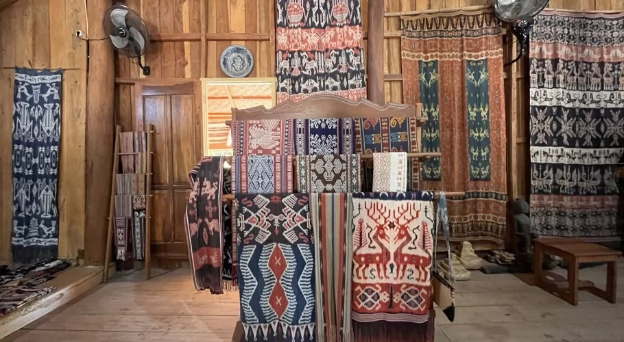 Numerous vibrant ikat textiles with intricate patterns are displayed on wooden racks and walls inside a warmly lit wooden gallery.