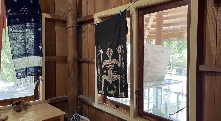 Inside a wooden gallery, two handcrafted ikat textiles hang in sunlit windows; one is dark blue and white, the other black with a detailed figure.