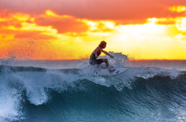 A surfer with long blonde hair rides a breaking wave, creating white spray against a vibrant orange and yellow sunset sky over the ocean.