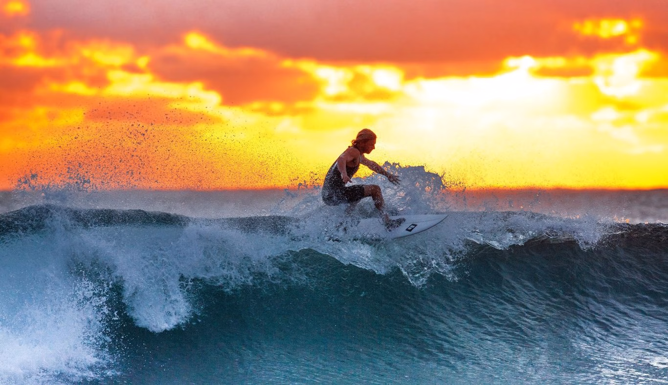 A surfer with long blonde hair rides a breaking wave, creating white spray against a vibrant orange and yellow sunset sky over the ocean.