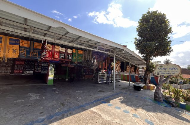 Katonga Humba, an open-air shop in Sumba, displays numerous colorful Ikat textiles and traditional accessories under a metal roof on a sunny day.