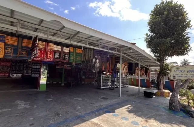 Katonga Humba, an open-air shop in Sumba, displays numerous colorful Ikat textiles and traditional accessories under a metal roof on a sunny day.