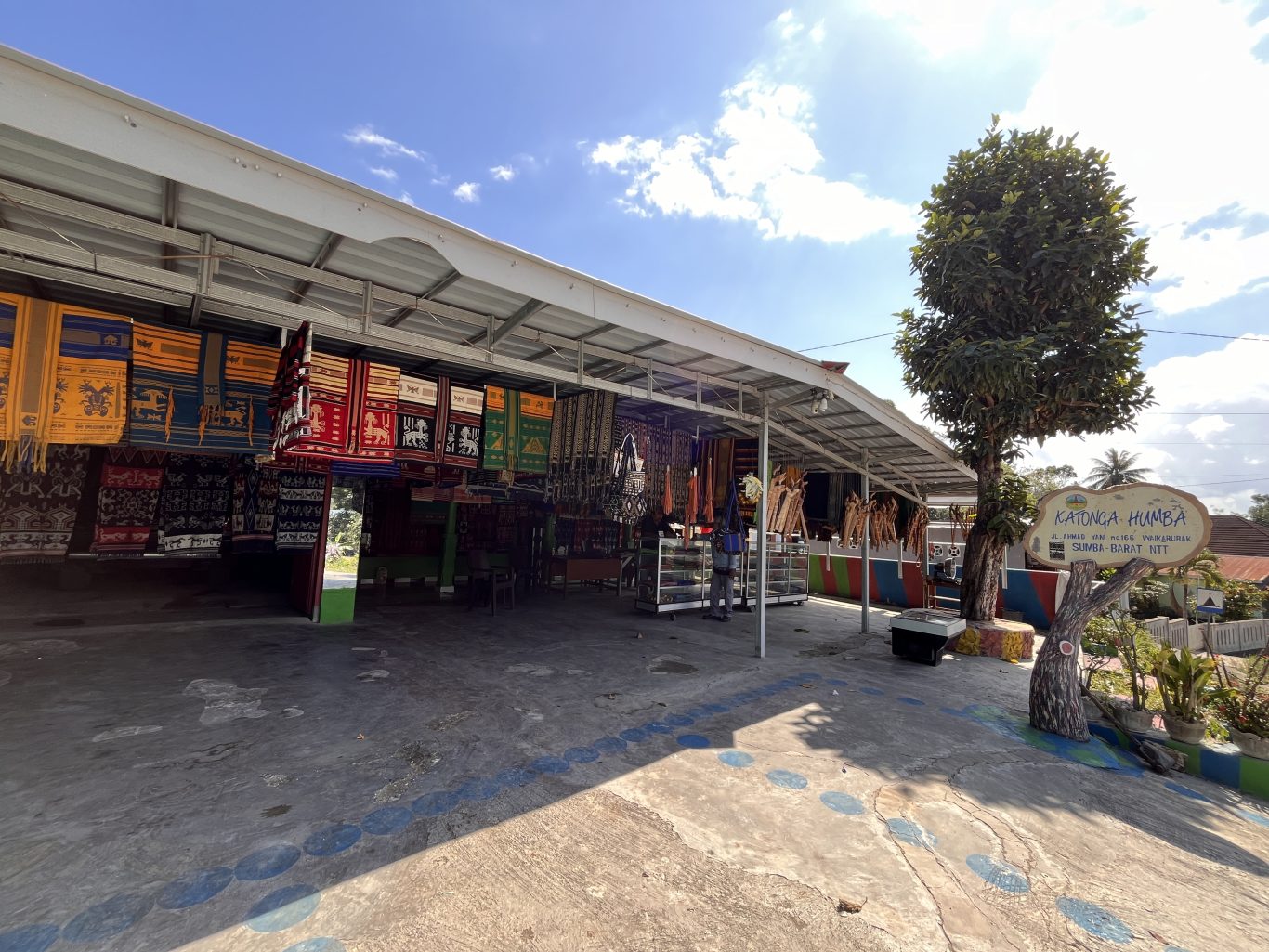 Katonga Humba, an open-air shop in Sumba, displays numerous colorful Ikat textiles and traditional accessories under a metal roof on a sunny day.