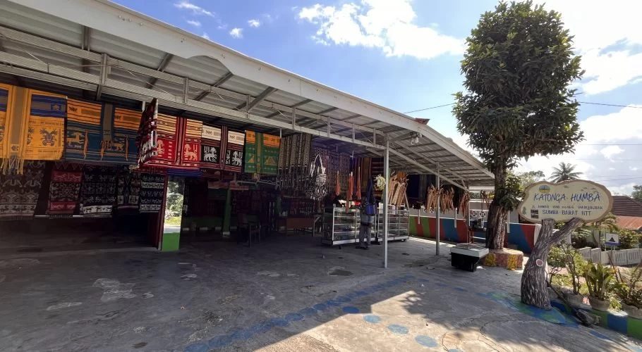 Katonga Humba, an open-air shop in Sumba, displays numerous colorful Ikat textiles and traditional accessories under a metal roof on a sunny day.