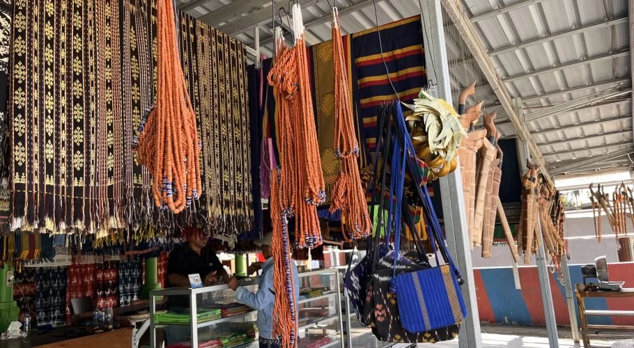 Inside a Sumba souvenir shop, dark patterned ikat textiles and long orange beaded necklaces hang from the ceiling, with wooden carvings and people visible.