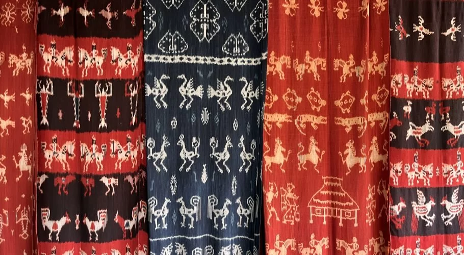 Several vibrant Sumba Ikat textiles hang vertically, showcasing intricate patterns of horsemen, birds, and traditional motifs in red, black, white, and dark blue.