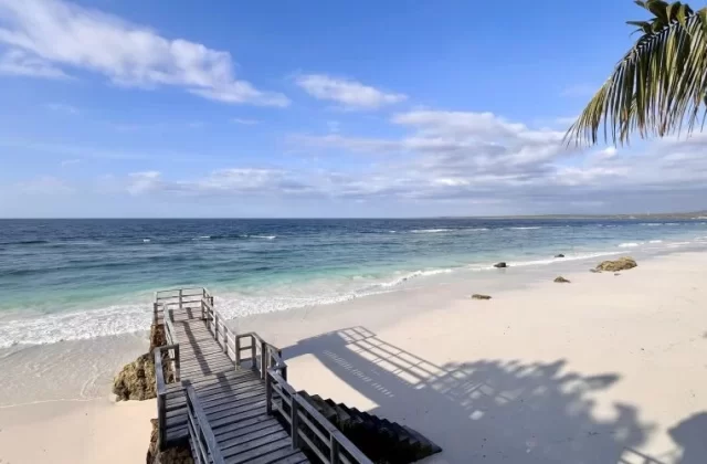A wooden walkway descends to a pristine white sand beach with clear turquoise waters under a blue sky, framed by palm fronds and their shadows.