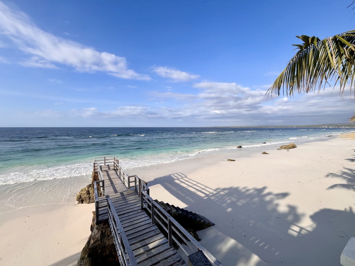 A wooden walkway descends to a pristine white sand beach with clear turquoise waters under a blue sky, framed by palm fronds and their shadows.