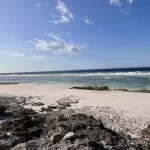 A wide white sand beach with dark rocky foreground meets turquoise and dark blue ocean waters under a bright blue sky with scattered clouds.