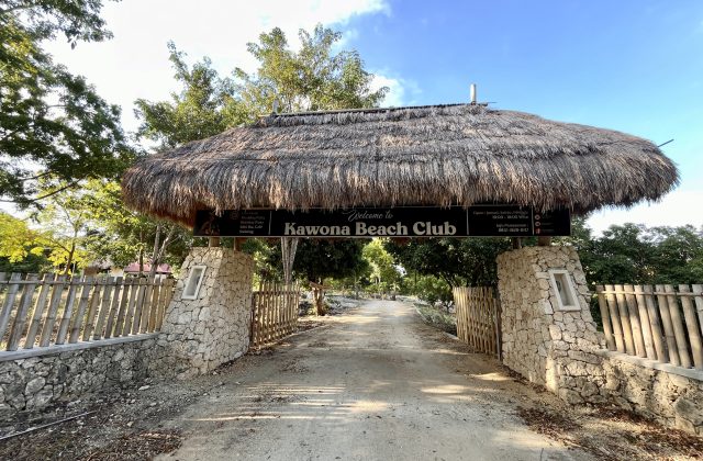 A thatched roof entrance sign for Kawona Beach Club stands over a dirt road, flanked by stone pillars and bamboo fences under a clear blue sky.
