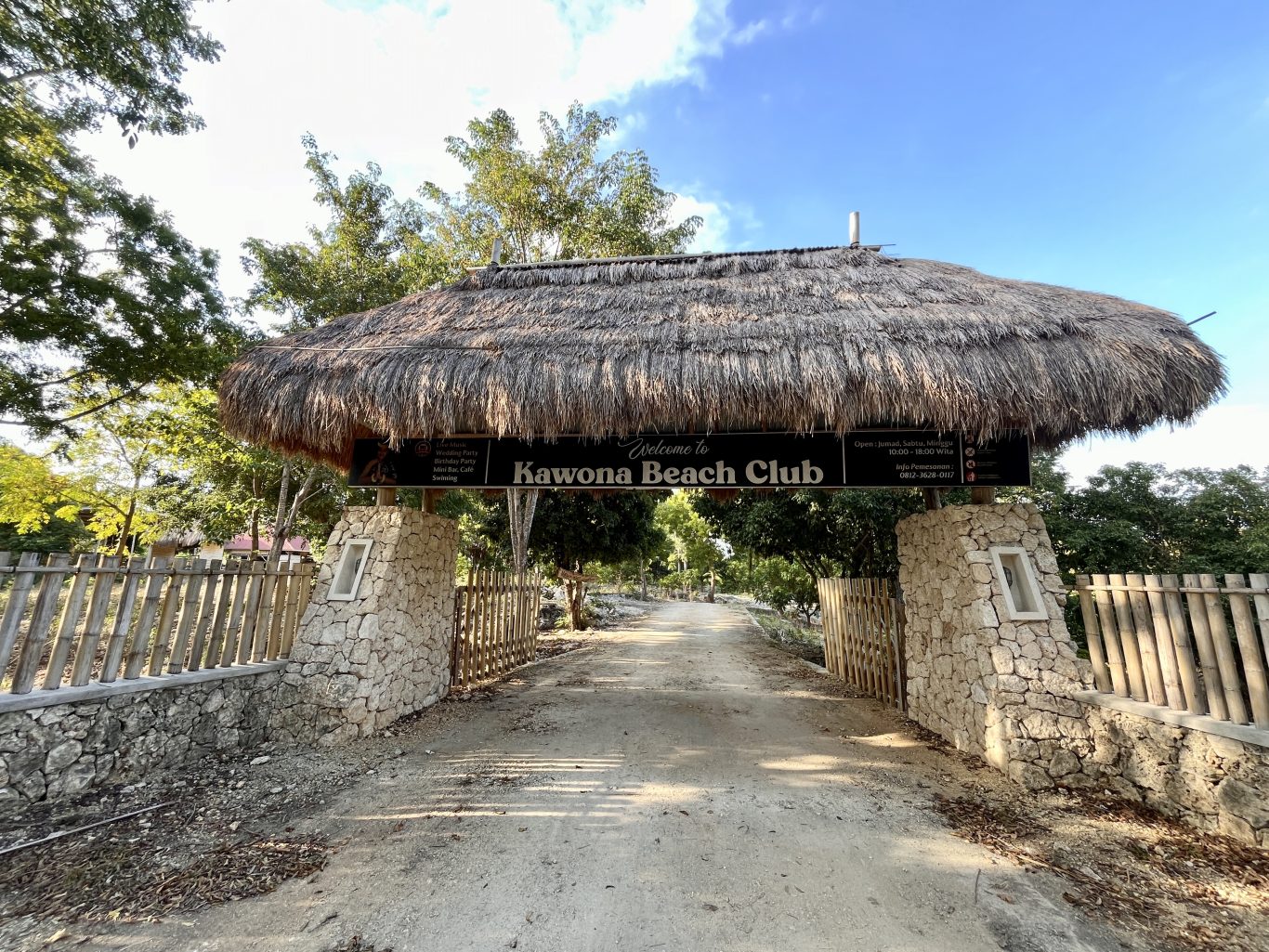 A thatched roof entrance sign for Kawona Beach Club stands over a dirt road, flanked by stone pillars and bamboo fences under a clear blue sky.