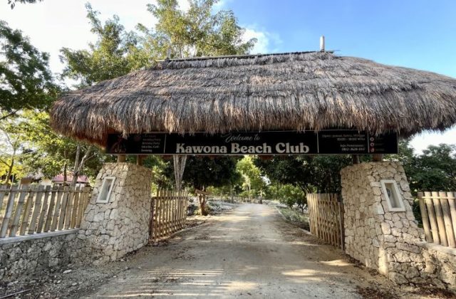 A thatched roof entrance sign for Kawona Beach Club stands over a dirt road, flanked by stone pillars and bamboo fences under a clear blue sky.