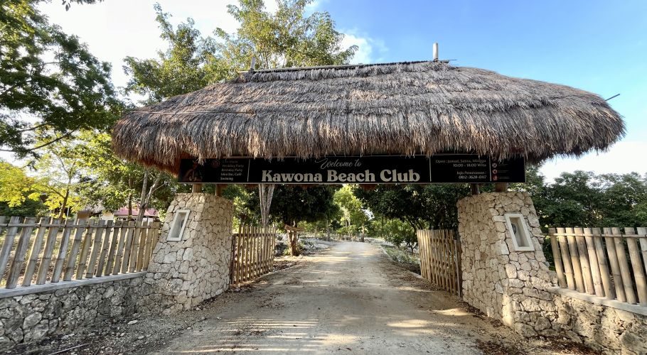 A thatched roof entrance sign for Kawona Beach Club stands over a dirt road, flanked by stone pillars and bamboo fences under a clear blue sky.