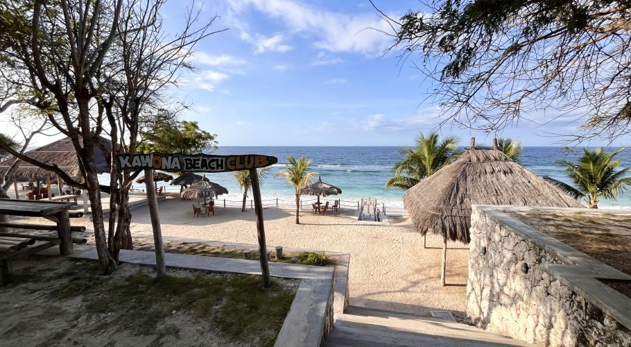Kawona Beach Club on a white sand beach features thatched huts, palm trees, and turquoise ocean water under a blue sky.