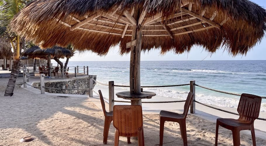 A thatched umbrella shades brown chairs on a white sandy beach next to a blue ocean with gentle waves under a clear sky.