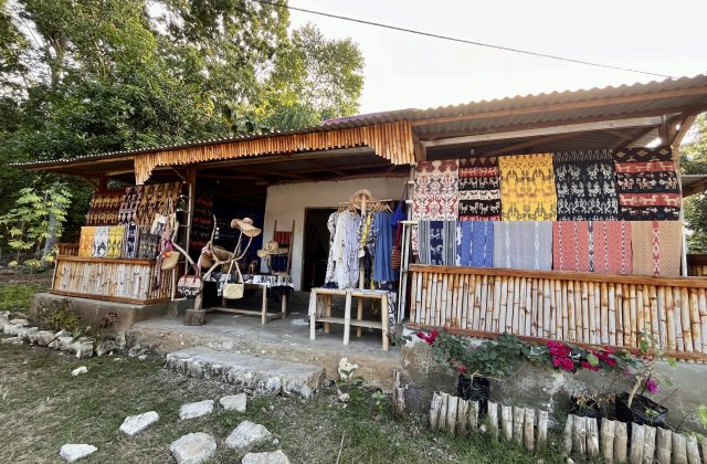 A souvenir shop with a corrugated roof and bamboo railing displays colorful Sumba ikat textiles, clothing, hats, and bags. Green trees are in the background, with pink flowers in the foreground.