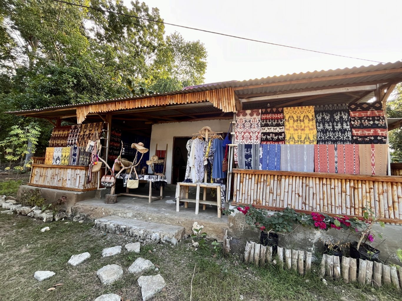 A souvenir shop with a corrugated roof and bamboo railing displays colorful Sumba ikat textiles, clothing, hats, and bags. Green trees are in the background, with pink flowers in the foreground.