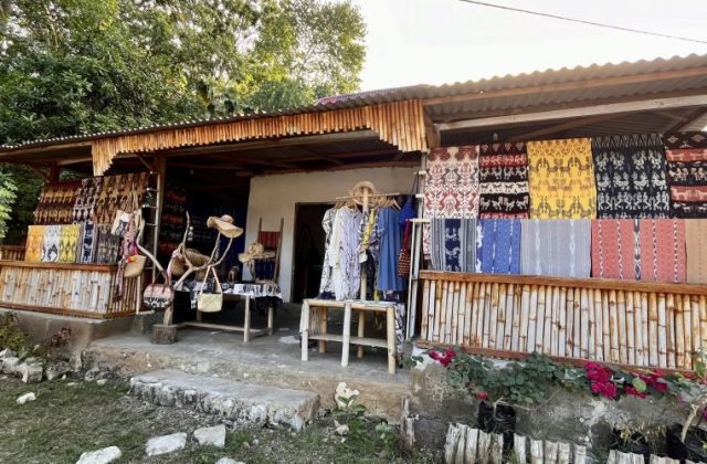 A souvenir shop with a corrugated roof and bamboo railing displays colorful Sumba ikat textiles, clothing, hats, and bags. Green trees are in the background, with pink flowers in the foreground.